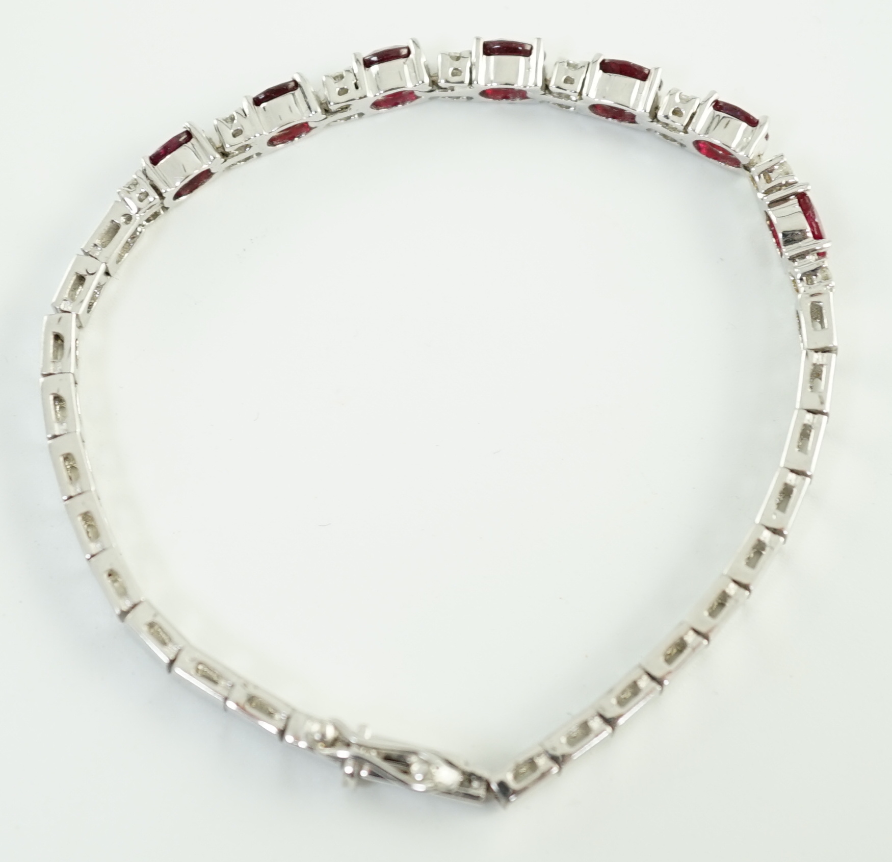 A 750 white gold and seven stone oval cut ruby set line bracelet, set with sixteen stone diamond spacers